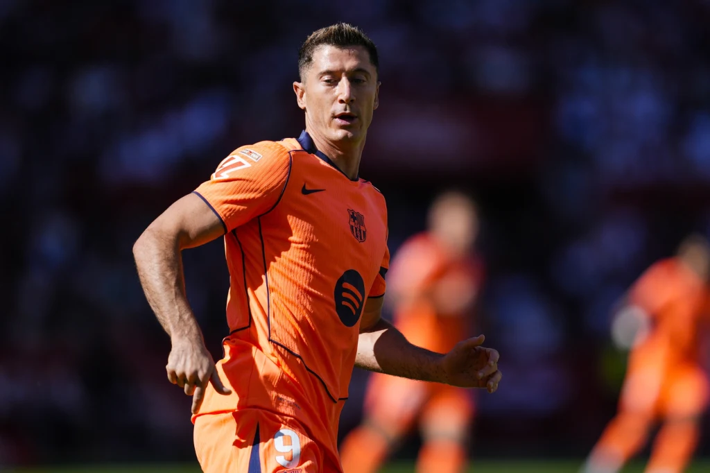 Piłkarz w pomarańczowym stroju sportowym z logo klubu, ujęty podczas gry na boisku, w tle rozmyta publiczność i pozostali zawodnicy.