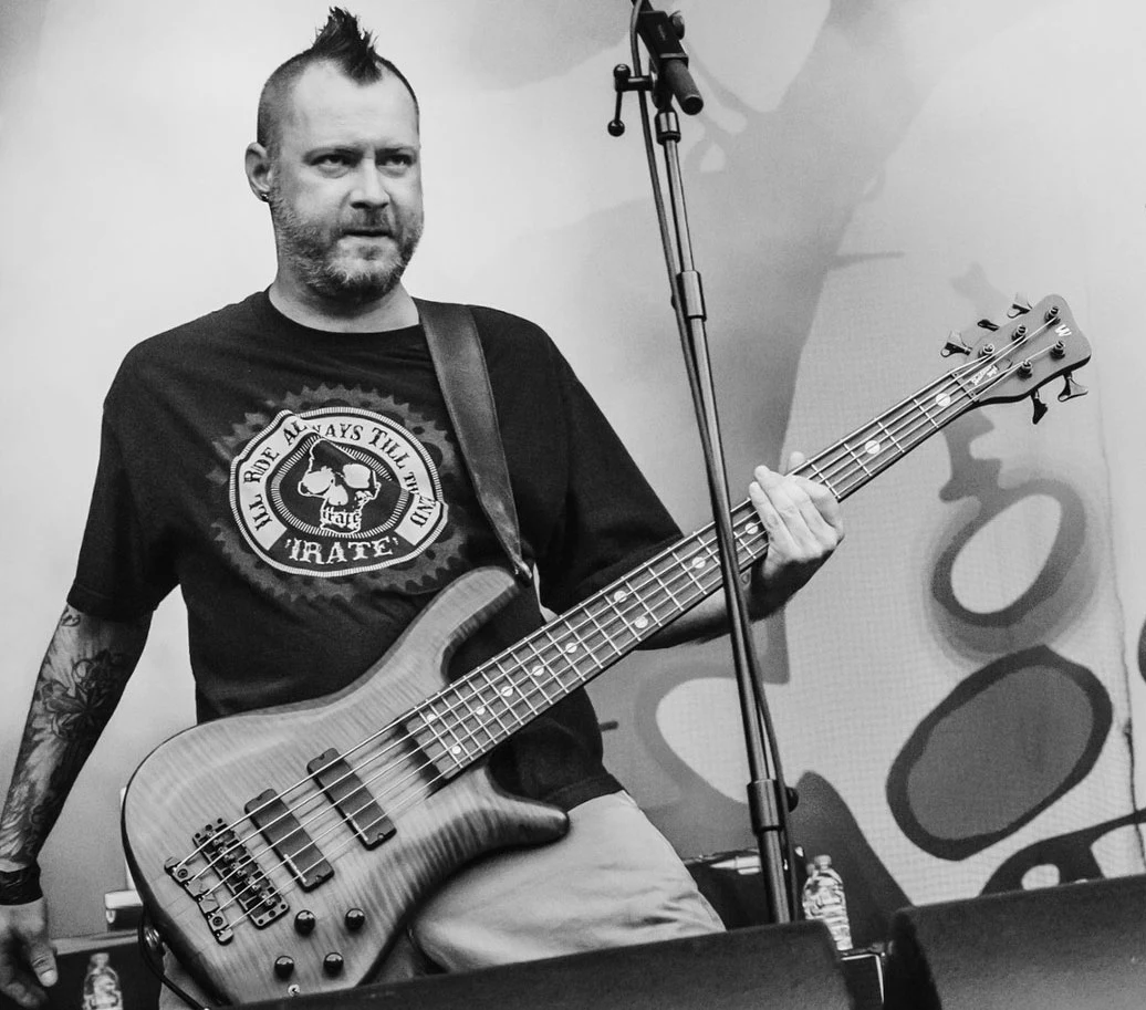 Sam Rivers (Limp Bizkit) miał 48 lat Mężczyzna z irokezem i tatuażami gra na gitarze basowej na koncercie, w tle widoczny jest mikrofon, zarys sceny oraz fragmenty sprzętu muzycznego.
