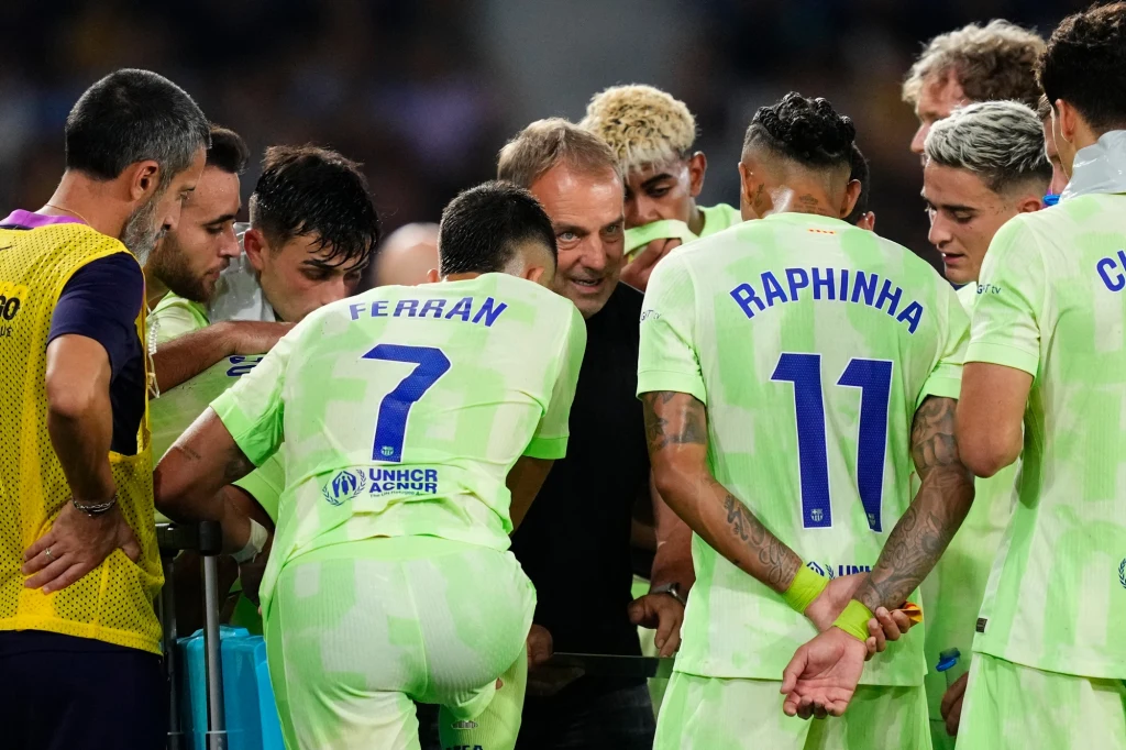 Hansi Flick z piłkarzami FC Barcelony Grupa piłkarzy ubranych w jasnozielone stroje stoi w półkolu wokół trenera, uważnie słuchając jego wskazówek. Na plecach zawodników widoczne są nazwiska i numery, atmosfera skupienia i zaangażowania.