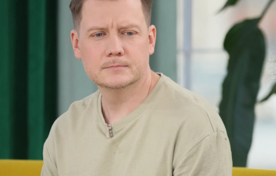 Jakub Tolak Mężczyzna o jasnej cerze i krótkich włosach w jasnej koszulce z zamkiem patrzy przed siebie z poważnym wyrazem twarzy, siedzi na żółtej kanapie, w tle duża roślina doniczkowa i zielona zasłona.