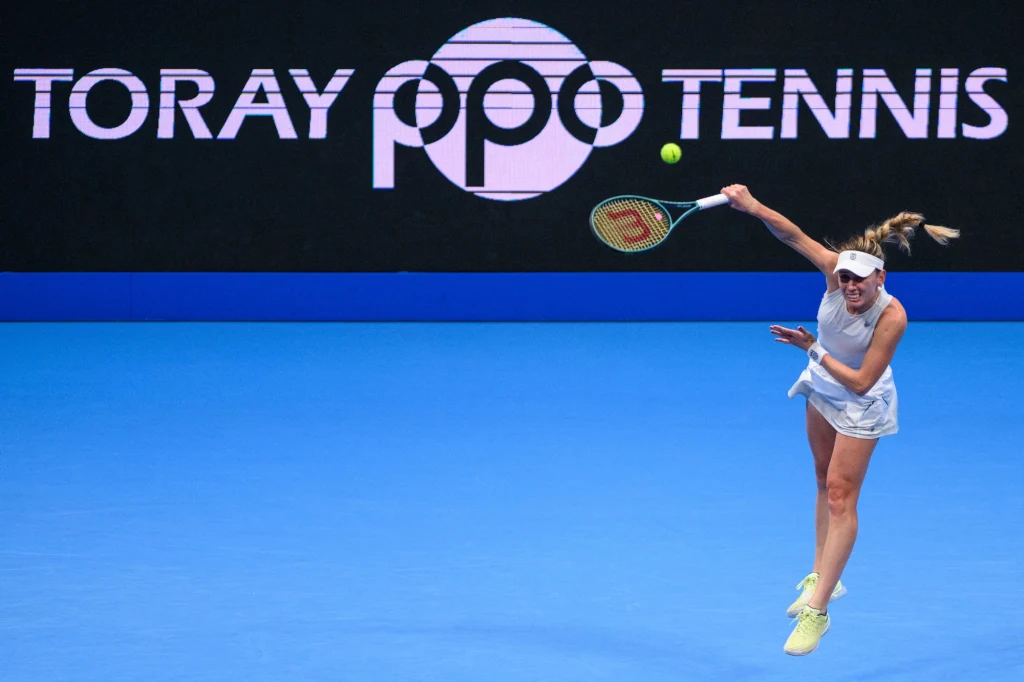 Tenisistka wykonująca dynamiczny serwis na niebieskim korcie, w tle biały napis TORAY PPO TENNIS na czarnym tle, piłka tenisowa unosząca się w powietrzu.