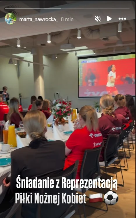 Grupa kobiet ubranych w sportowe koszulki siedzi przy wspólnym stole, na którym znajdują się śniadaniowe potrawy, napoje i kwiaty. W tle na dużym ekranie wyświetlany jest mecz piłki nożnej kobiet.