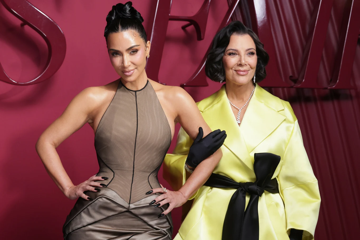 Fani twierdzą, że Kris Jenner wyglądem przypomina już własne córki Dwie elegancko ubrane kobiety pozujące razem na tle bordowej ściany, jedna z nich w dopasowanej sukni w odcieniach szarości, druga w żółtym płaszczu przewiązanym czarnym paskiem. Obie uśmiechnięte, w stylowych fryzurach, podkreślają modowy charakter sc...