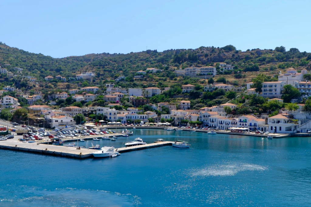 Malowniczy port z cumującymi jachtami i łodziami otoczony białymi budynkami o niebieskich okiennicach, położony u podnóża wzgórz pokrytych zielenią. W tle wzgórza z zabudowaniami, niebo bezchmurne.