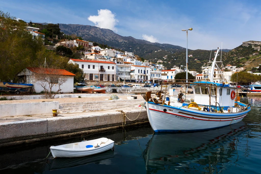 Biała łódź rybacka przycumowana do nabrzeża w malowniczym porcie, w tle niskie zabudowania miasteczka przy stromym zboczu gór, spokojna woda i przejrzyste niebo z pojedynczym obłokiem.
