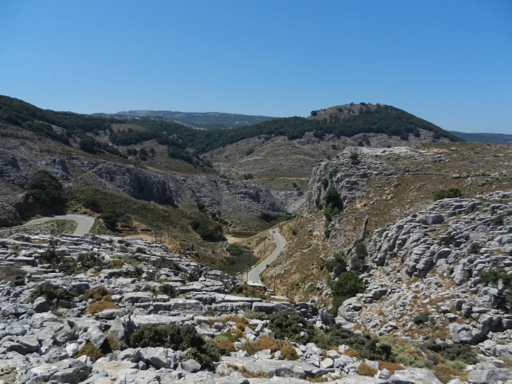 Krajobraz górski z licznymi kamieniami, skalistymi wzgórzami i wijącą się drogą przecinającą teren. Roślinność jest rzadka, występują głównie krzewy i niskie drzewa. W tle widoczne są zielone wzgórza, pod błękitnym niebem bez chmur.