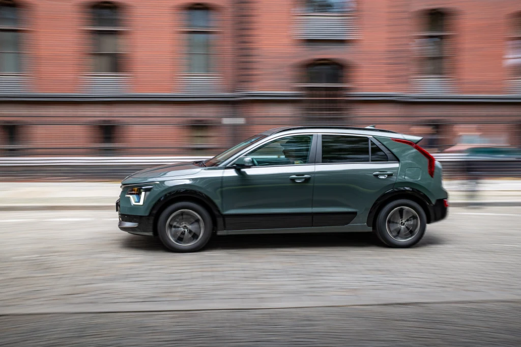 Samochód osobowy typu SUV w kolorze ciemnozielonym porusza się dynamicznie miejską ulicą, w tle znajduje się zabytkowy budynek z czerwonej cegły z dużymi oknami.