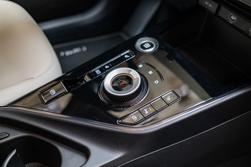 Nowoczesna konsola środkowa samochodu z elektroniczną gałką zmiany biegów, przyciskami sterującymi funkcjami pojazdu, czarnym wykończeniem oraz fragmentem jasnego fotela pasażera.
