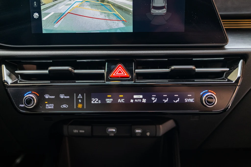 Panel klimatyzacji i systemu multimedialnego w samochodzie z wyświetlaczem prezentującym widok z kamery cofania oraz przycisk awaryjnych świateł trwałych pośrodku.