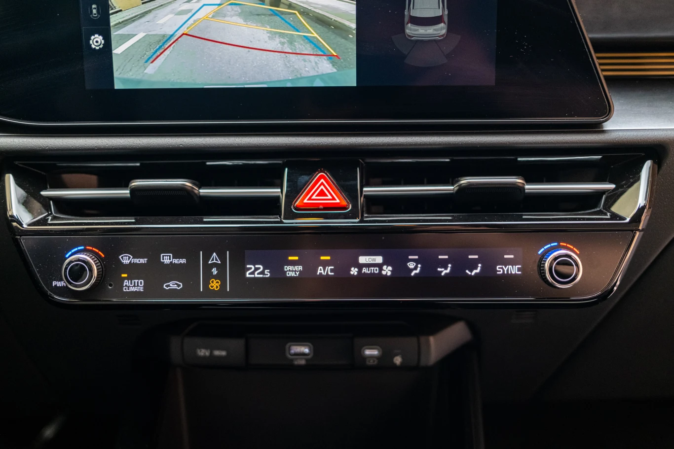 Panel klimatyzacji i systemu multimedialnego w samochodzie z wyświetlaczem prezentującym widok z kamery cofania oraz przycisk awaryjnych świateł trwałych pośrodku.
