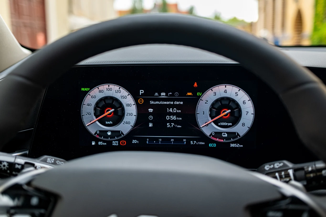 Cyfrowy panel wskaźników samochodowych z widocznymi prędkościomierzem, obrotomierzem oraz ekranem wyświetlającym aktualne dane pojazdu, takie jak prędkość, przebieg oraz zużycie paliwa, uchwycone zza kierownicy.