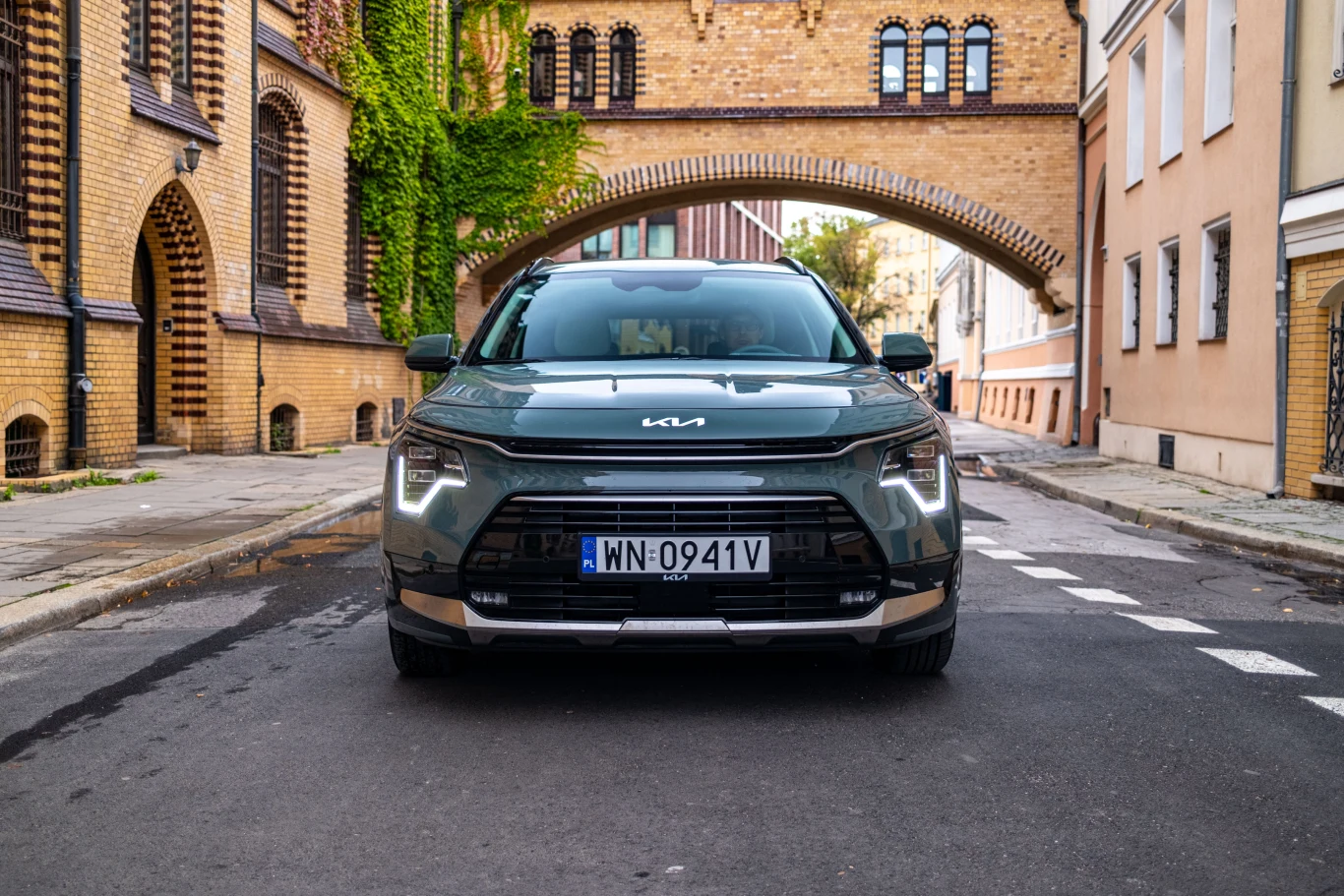 Nowoczesny zielony samochód Kia Niro ustawiony na środku wąskiej, brukowanej ulicy otoczonej zabytkowymi budynkami z czerwonej cegły i jasnego tynku. W tle charakterystyczny łuk łączący budynki, dodający klimatu starego miasta.