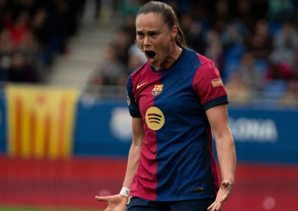 Ewa Pajor Piłkarka drużyny FC Barcelona w trakcie meczu, wyrażająca silne emocje, stojąca na tle boiska sportowego. Kobieta w koszulce klubowej z szeroko otwartymi ustami i zaciśniętymi pięściami, w tle rozmyte sylwetki kibiców i elementy stadionu.