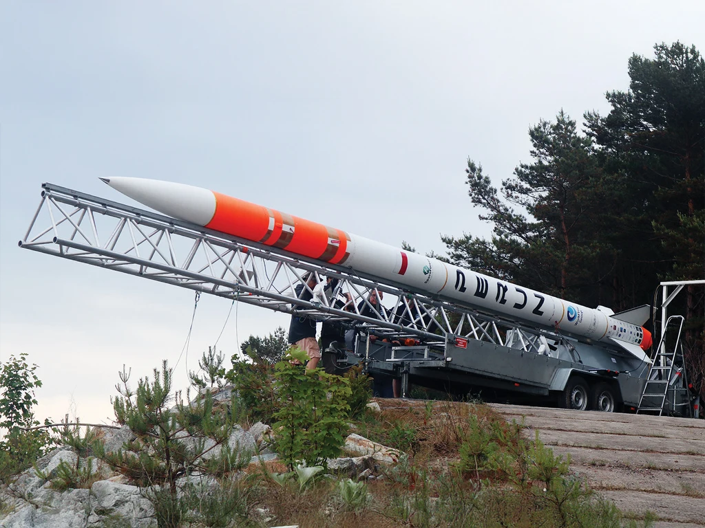 Rakieta Perun na poligonie Sił Powietrznych w Ustce Rakieta o pomarańczowo-białym korpusie zamocowana na mobilnej platformie startowej wśród roślinności i leśnego otoczenia, techniczna infrastruktura oraz fragmenty drzew w tle.