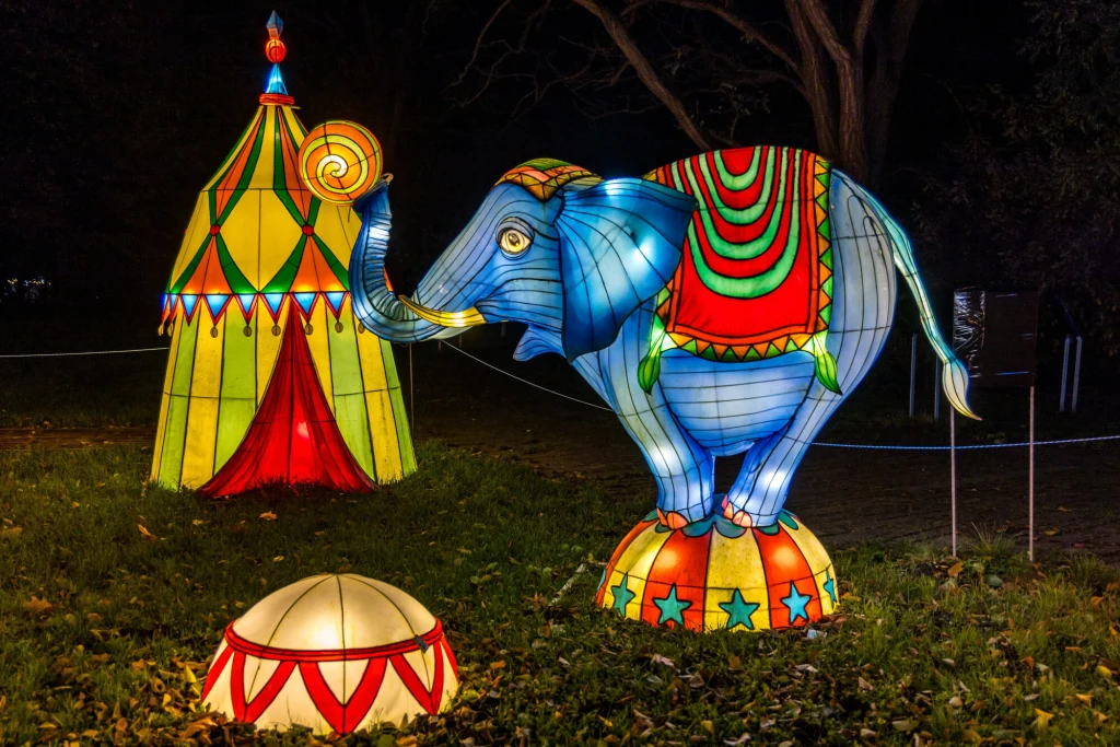 Kolorowy, podświetlony słoń stojący na kulistym podeście, ubrany w ozdobne szaty, obok namiot w stylu cyrkowym oraz inne świetlne dekoracje na tle nocnego parku.