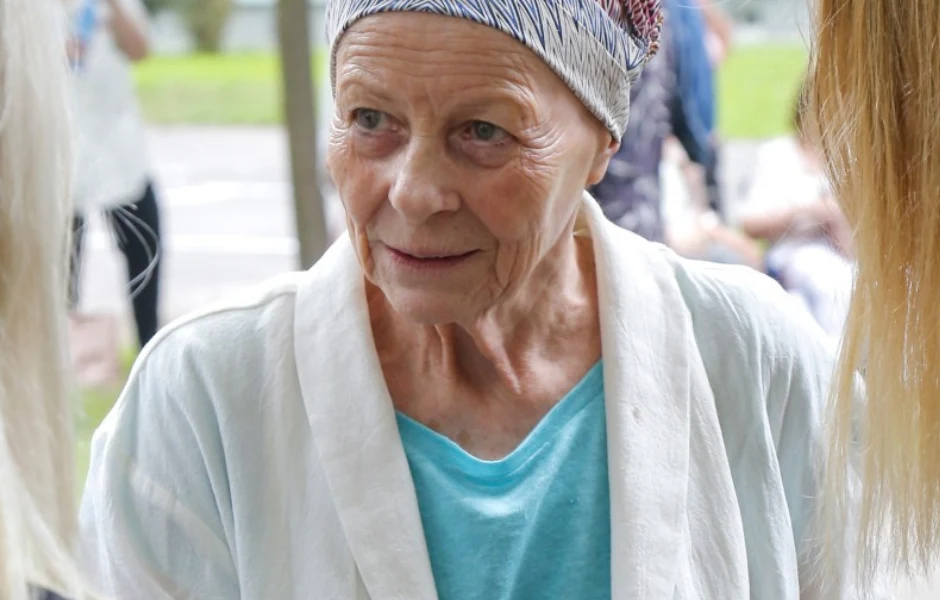 Anna Nehrebecka Starsza kobieta z opaską na głowie i uśmiechem na twarzy, ubrana w jasny szlafrok i niebieską koszulkę, rozmawia z innymi osobami na tle rozmytego parku.