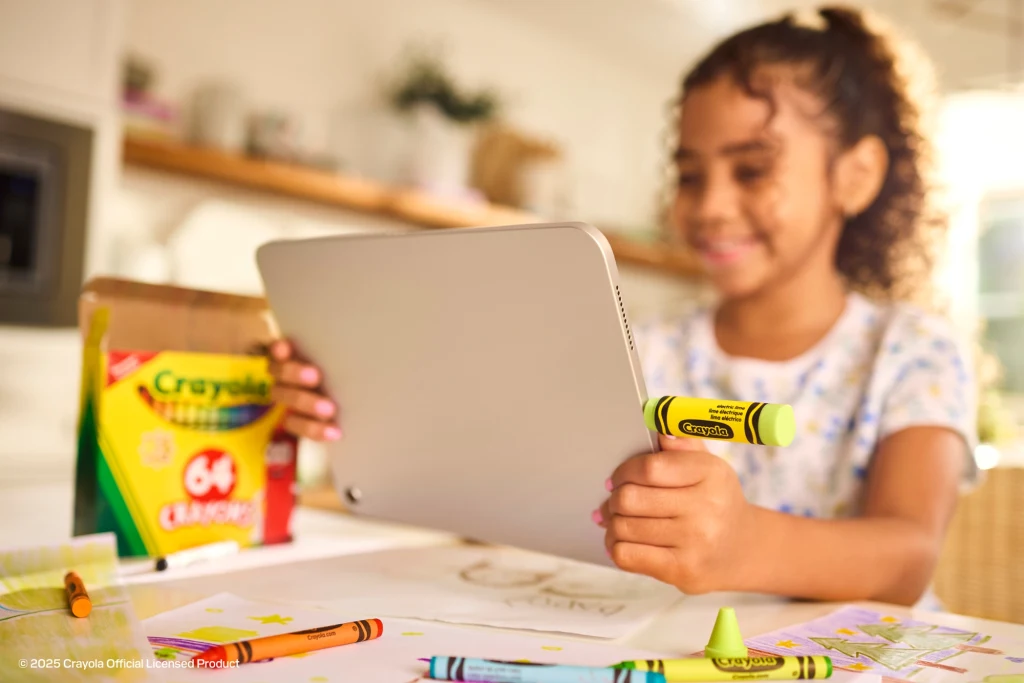 Sandisk Crayola USB-C Flash Drive to pendrive w kształcie kredki. Dziewczynka z uśmiechem trzyma tablet, otoczona kolorowymi kredkami Crayola i kartkami z rysunkami. Na biurku pudełko kredek oraz rozrzucone przybory plastyczne, w tle domowe wnętrze.