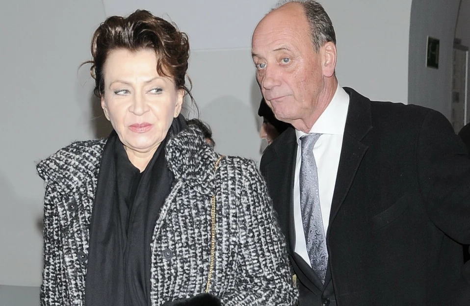 Grażyna Strachota i Dariusz Szpakowski. Kobieta o ciemnych, upiętych włosach w eleganckim płaszczu w kratę i szaliku stoi obok mężczyzny w garniturze i krawacie, oboje mają poważne miny, tło jasne i neutralne.