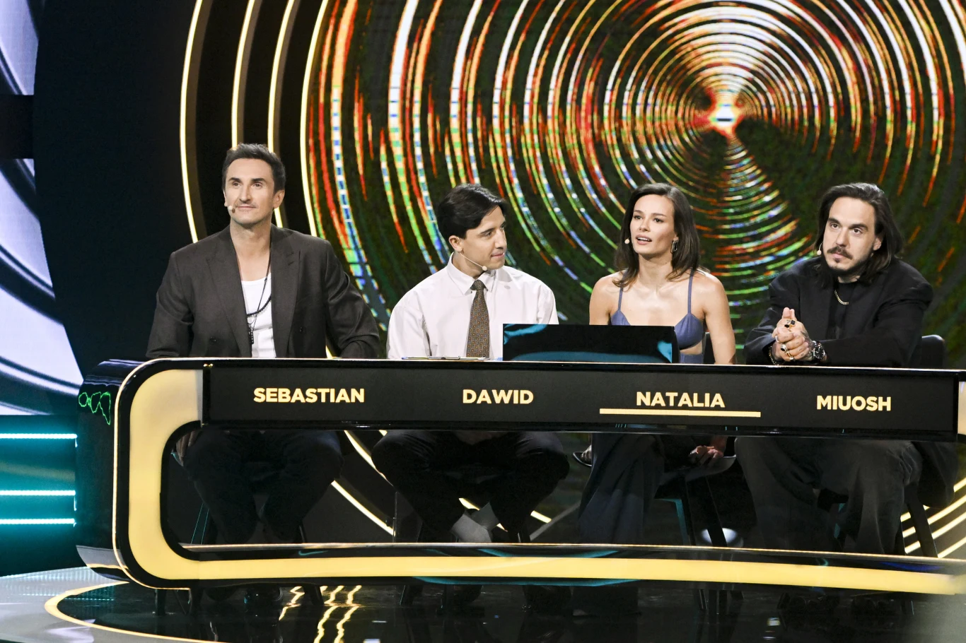 Sebastian Karpiel-Bułecka (Zakopower) zaprosił swoich kolegów z jury "Must Be The Music": Dawida Kwiatkowskiego, Natalię Szroeder i Miuosha Czwórka jurorów siedzi za stołem z podświetlonymi imionami na froncie, w tle efektowna grafika przypominająca wirującą spiralę.
