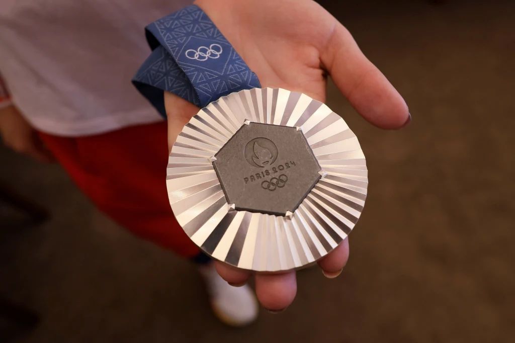 Srebrny medal olimpijski z logo Igrzysk Olimpijskich w Paryżu 2024 trzymany w wyciągniętej dłoni. Niebieska wstążka z symbolem olimpijskim przymocowana do medalu, w tle widoczna fragmentarycznie ręka i ubranie osoby.