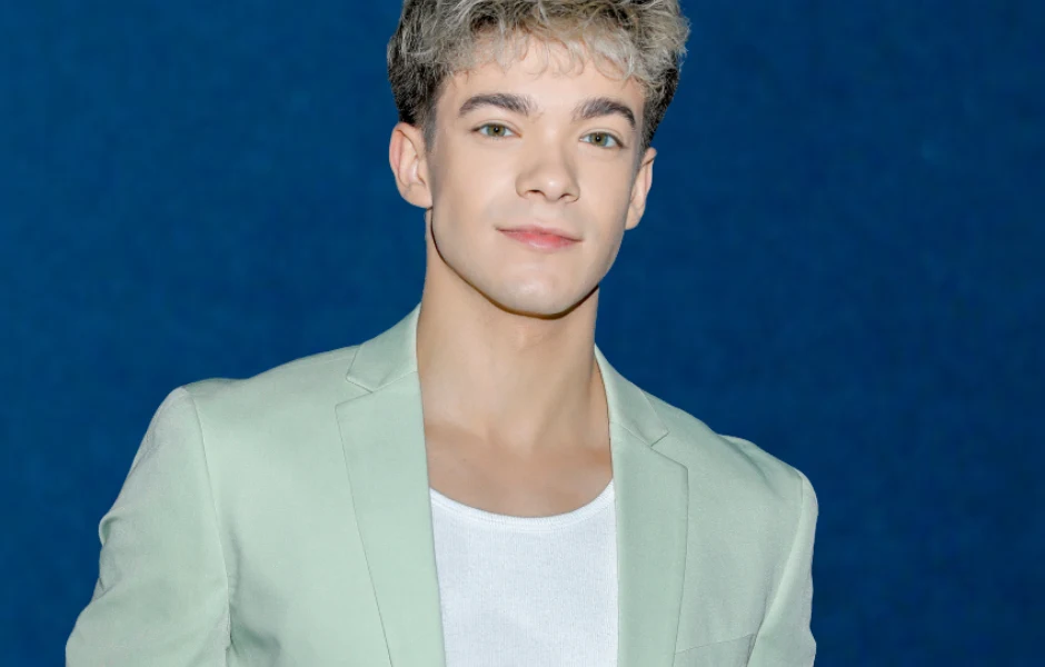 Kuba Szmajkowski Młody mężczyzna z jasnymi włosami ubrany w jasną marynarkę i biały podkoszulek na tle granatowej ściany.