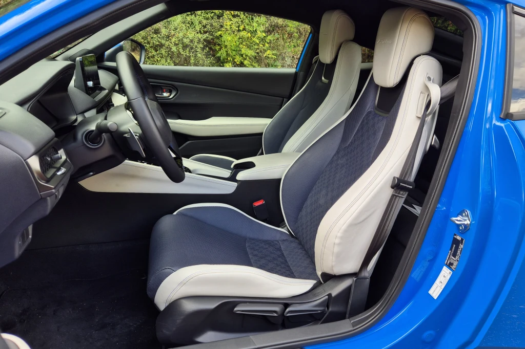 Fotele w nowej Hondzie Prelude są wygodne i zapewniają niezłe trzymanie boczne. Co ciekawe - fotel kierowcy oferuje nieco lepsze podparcie w zakrętach, zaś fotel pasażera nieco większy komfort Wnętrze sportowego samochodu z dwoma fotelami o wyprofilowanym kształcie, tapicerowanymi w odcieniach czerni i szarości, deską rozdzielczą z nowoczesnym panelem i kierownicą, drzwi otwarte, karoseria lakierowana na niebiesko.