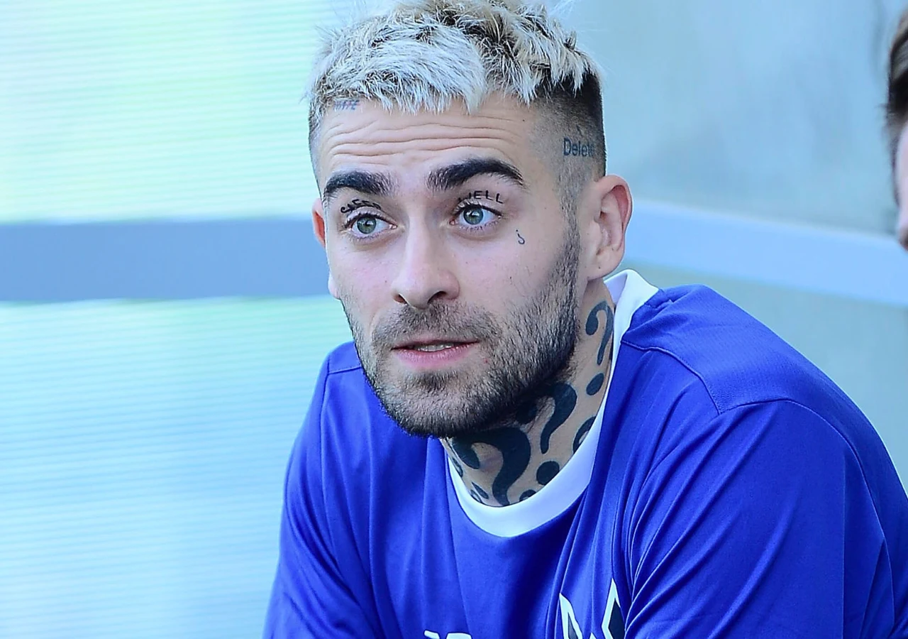 Mężczyzna z jasnymi, krótkimi włosami i tatuażami na twarzy oraz szyi, ubrany w niebieską sportową koszulkę, spogląda w bok z lekkim uśmiechem, siedząc w jasnym otoczeniu.