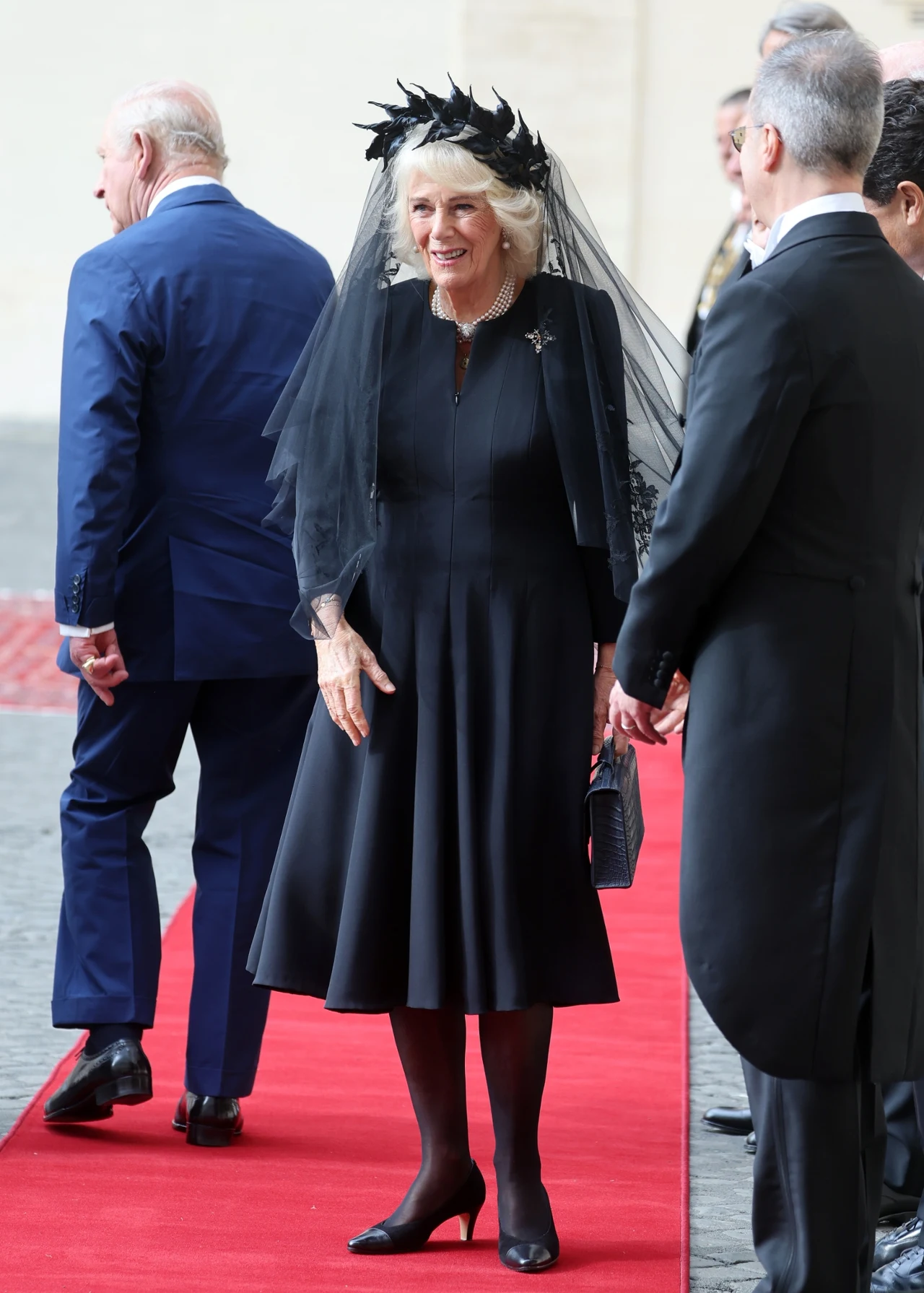 Dlaczego królowa Camilla musiała nosić czarną stylizację? Kobieta w czarnej, eleganckiej sukni i welonie, stojąca na czerwonym dywanie, otoczona przez kilku mężczyzn w garniturach, w tle widoczne są osoby odwrócone plecami.