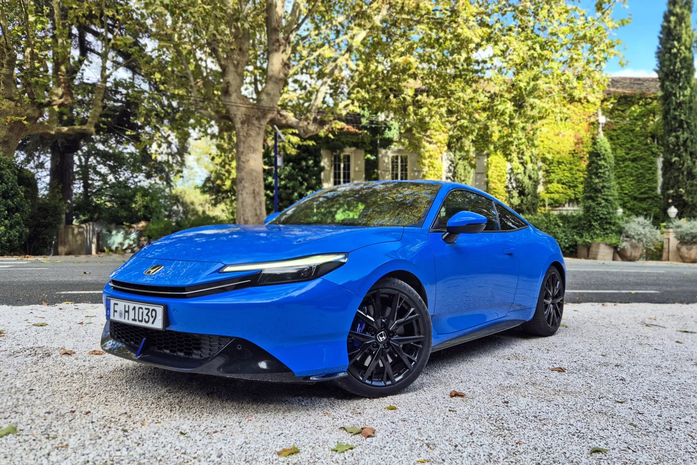 Honda Prelude powraca na rynek po 25 latach nieobecności Niepozorny, sportowy samochód osobowy w wyrazistym niebieskim kolorze zaparkowany na białym żwirze przy ulicy w spokojnej, zielonej okolicy, otoczony drzewami oraz krzewami, w tle widoczne budynki mieszkalne.