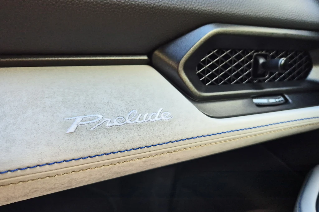 Skórzane wstawki z kontrastowym przeszyciem nadają wnętrzu Prelude nieco szlachetności Logo Prelude wyhaftowane na jasnej tapicerce wnętrza samochodu, obok kratki nawiewu o czarnej siateczkowej strukturze i przycisku, wykończone niebieskim przeszyciem.