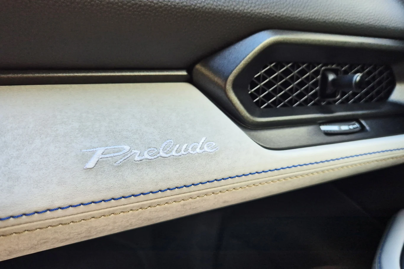 Skórzane wstawki z kontrastowym przeszyciem nadają wnętrzu Prelude nieco szlachetności Logo Prelude wyhaftowane na jasnej tapicerce wnętrza samochodu, obok kratki nawiewu o czarnej siateczkowej strukturze i przycisku, wykończone niebieskim przeszyciem.