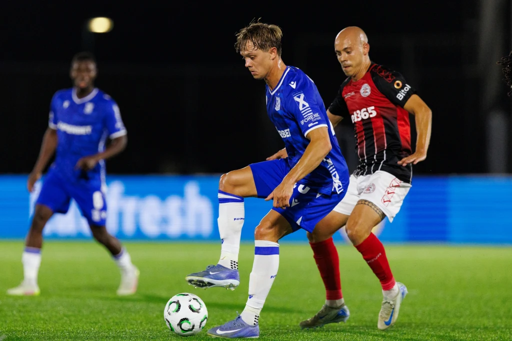 Lincoln Red Imps - Lech Poznań (23.10.2025) Dwóch piłkarzy rywalizuje o piłkę podczas meczu piłki nożnej na boisku, zawodnik w niebieskim stroju kontroluje piłkę, a piłkarz w czerwono-czarnym stroju zbliża się do niego, w tle widać innego gracza na rozmytym tle.