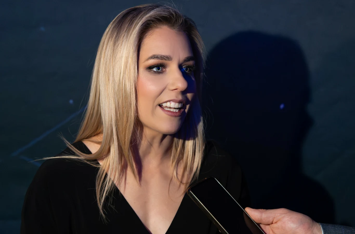 Kobieta z blond włosami w czarnym stroju udziela wywiadu. W tle widoczny cień jej sylwetki, obok pojawia się zbliżony mikrofon i dłoń osoby zadającej pytanie.