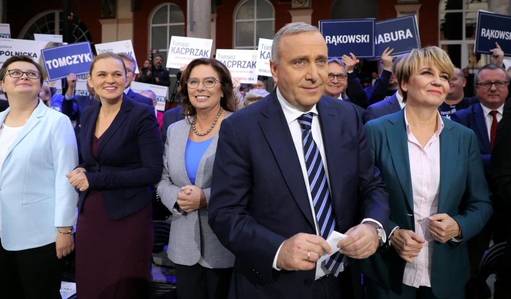 Grupa polityków uśmiechnięta, stojąca w pierwszym rzędzie podczas wiecu. W tle tłum z transparentami z nazwiskami kandydatów, otoczenie miejskie, atmosfera spotkania publicznego.