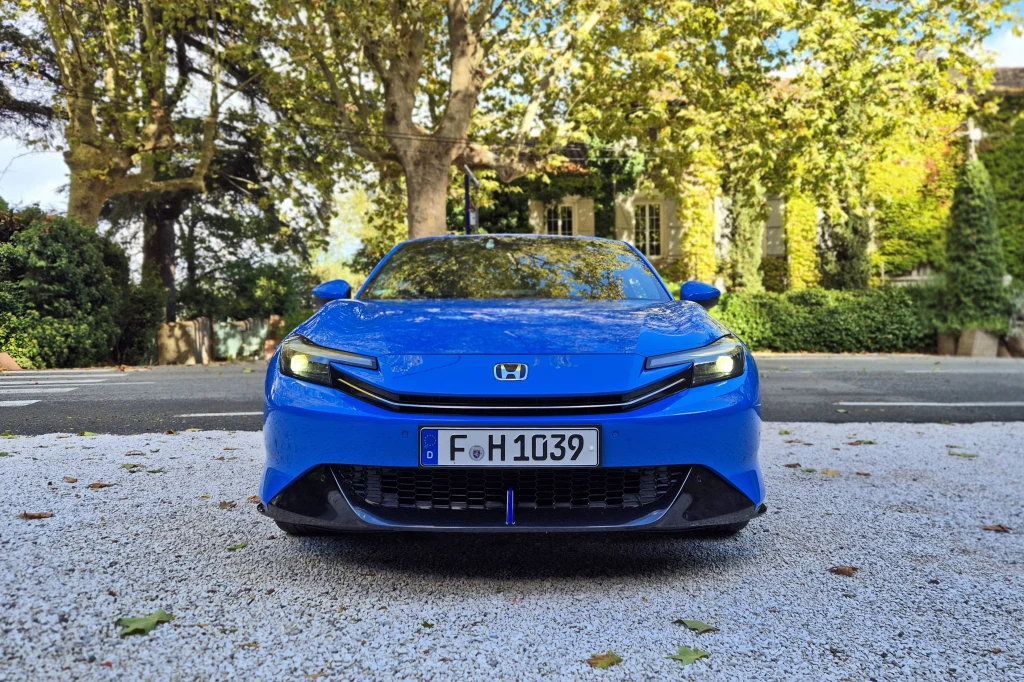 W dolną część zderzaka Prelude wkomponowano duży spojler, który generuje spory docisk podczas szybkiej jazdy Niebieski samochód osobowy marki Honda zaparkowany na jasnym żwirowym podłożu, ujęty od przodu, w tle gęste drzewa oraz budynek pokryty częściowo bluszczem.