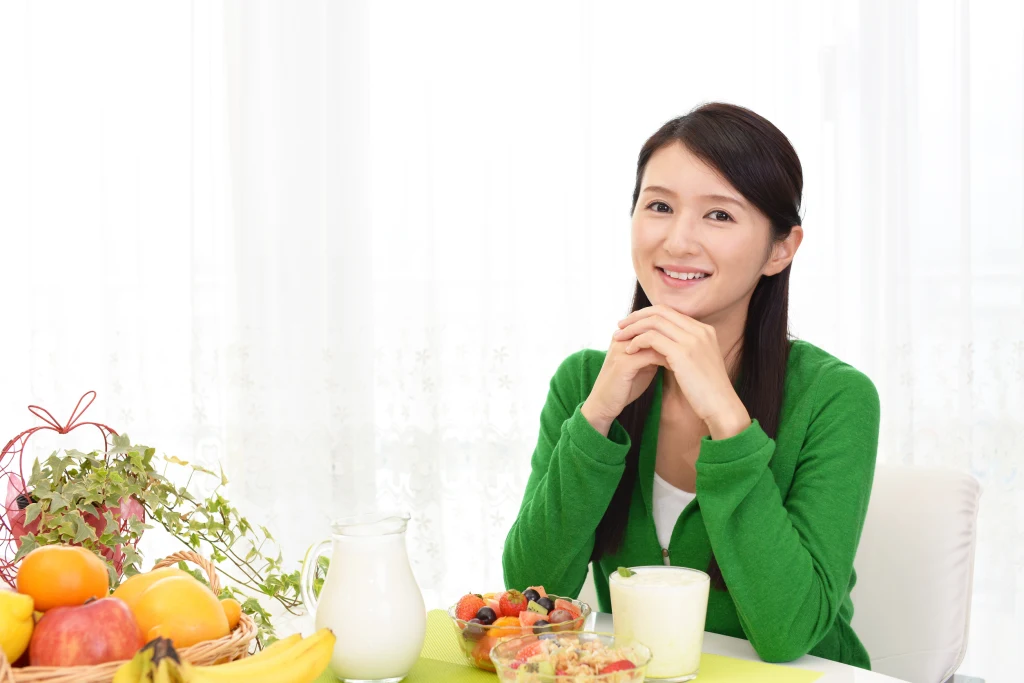 Wybór odpowiednich produktów pozwala tworzyć smaczny i zbilansowany jadłospis Młoda kobieta ubrana w zielony sweter siedzi przy stole zastawionym owocami, mlekiem oraz deserem, uśmiechając się w stronę aparatu.