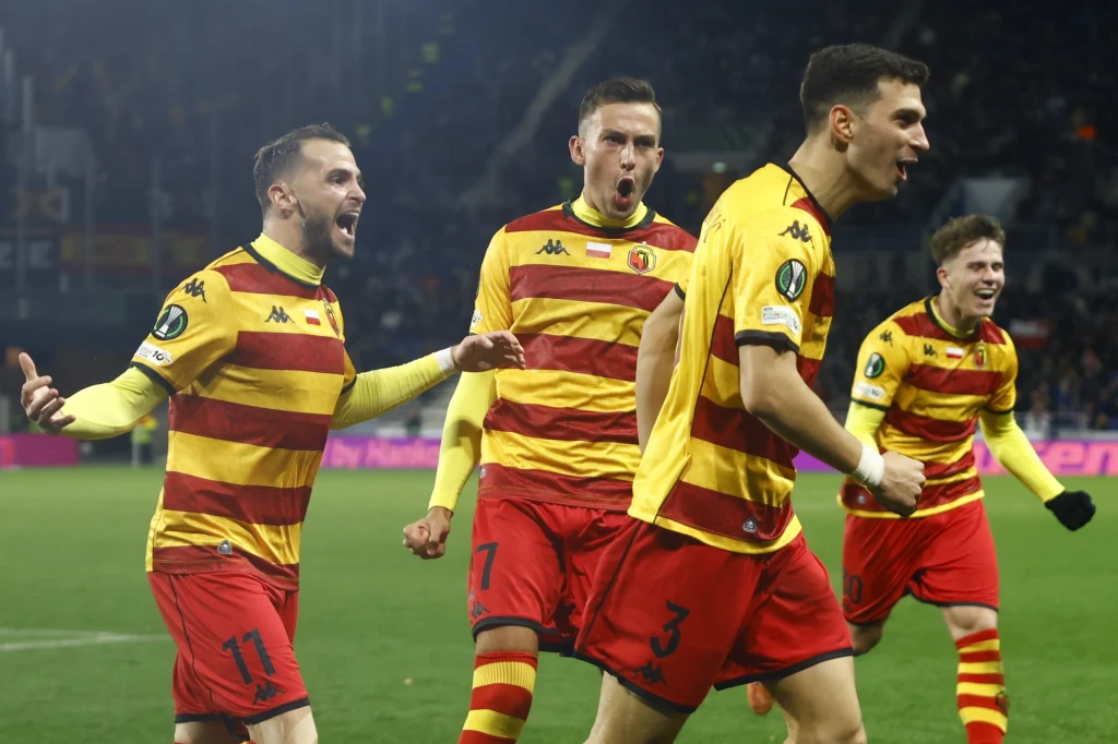 Piłkarze Jagielloni Białystok w sezonie 2025/2026 Czterech piłkarzy w żółto-czerwonych strojach świętuje zdobycie bramki na boisku, wyrażając radość i energię, tło rozmyte z widocznym stadionem oraz kibicami.