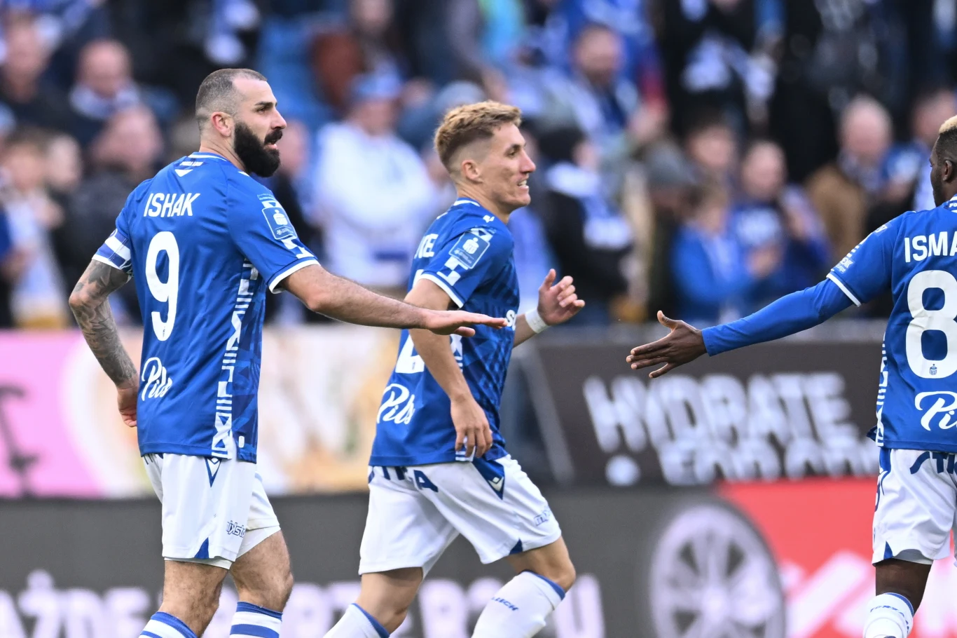 Piłkarze Lecha Poznań w meczu Ekstraklasy z Pogonią Szczecin Trzech piłkarzy drużyny w niebieskich koszulkach świętuje zdobycie bramki podczas meczu, otoczeni przez rozmyty tłum kibiców na trybunach.