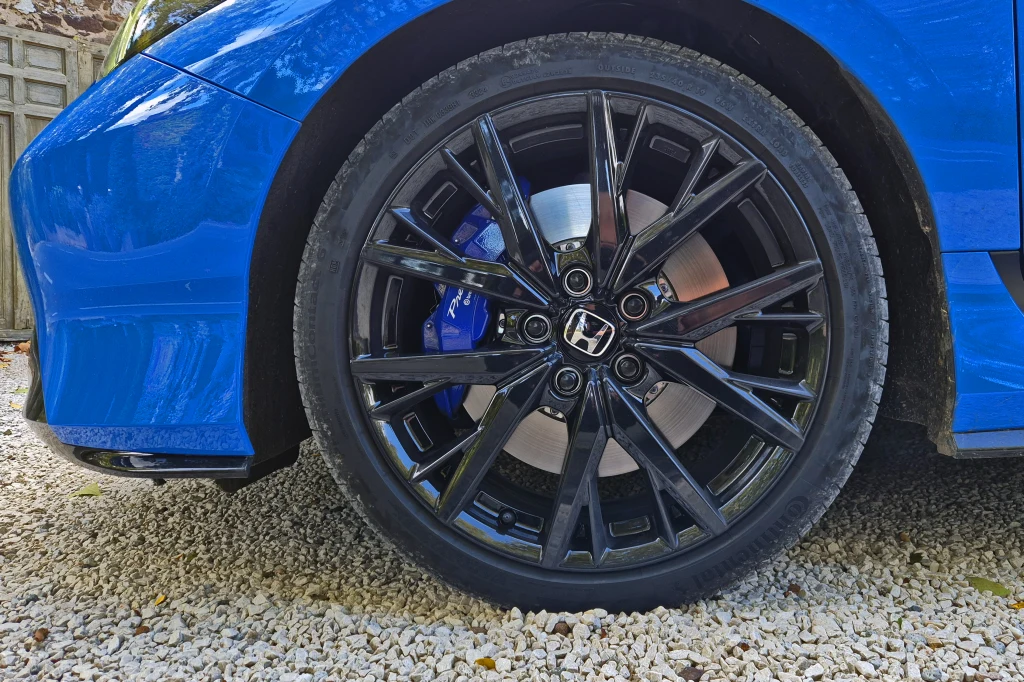 Prelude standardowo porusza się na 19-calowych felgach, ale opony mają rozmiar jedynie 235/40 Czarny, połyskujący felga aluminiowa z widocznym logo marki Honda na tle niebieskiego nadwozia samochodu i żwirowej nawierzchni.