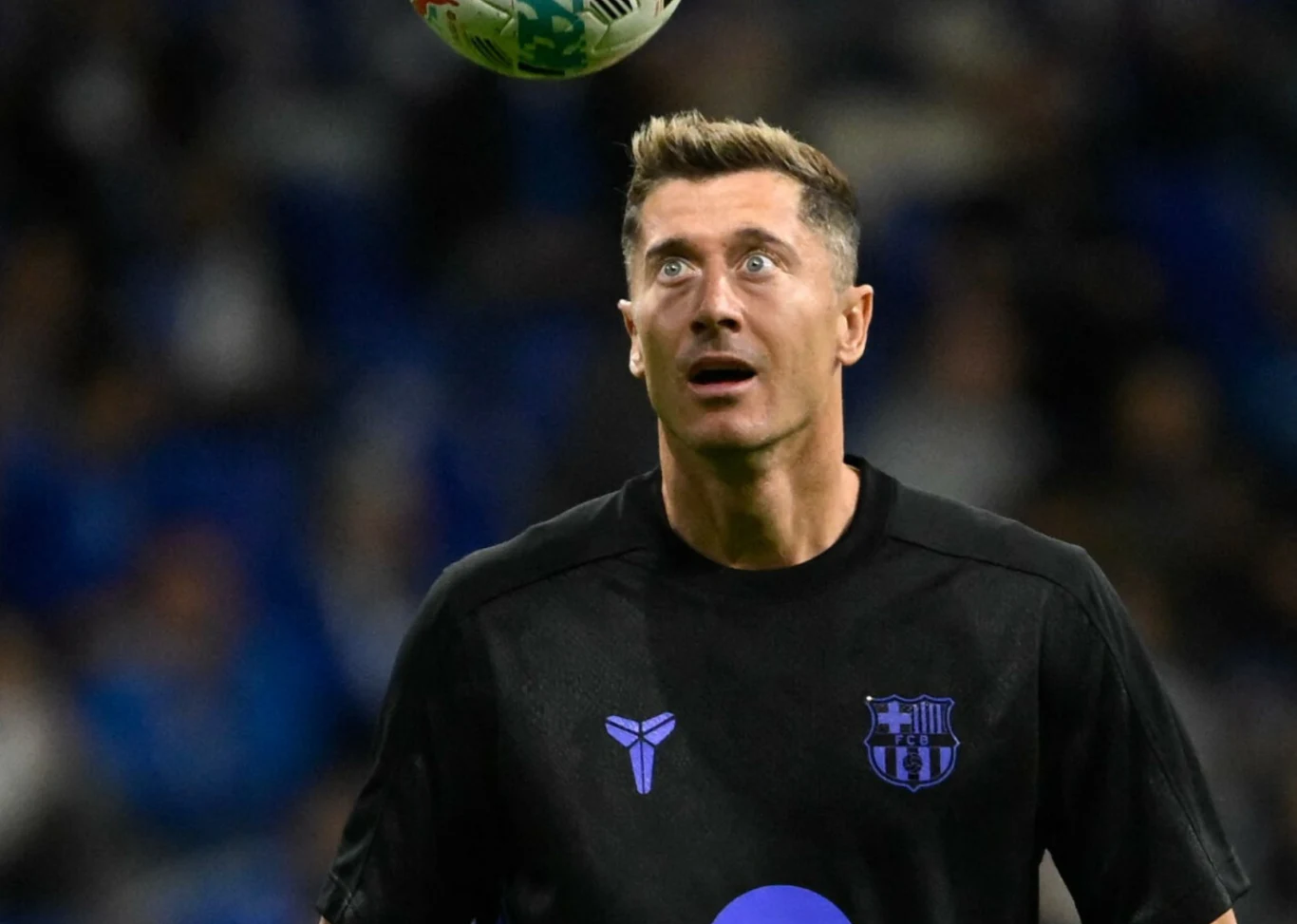 Piłkarz w czarnej koszulce Barcelony z wyraźnym skupieniem patrzy w górę, gdzie znajduje się piłka, na tle zamazanego stadionu.