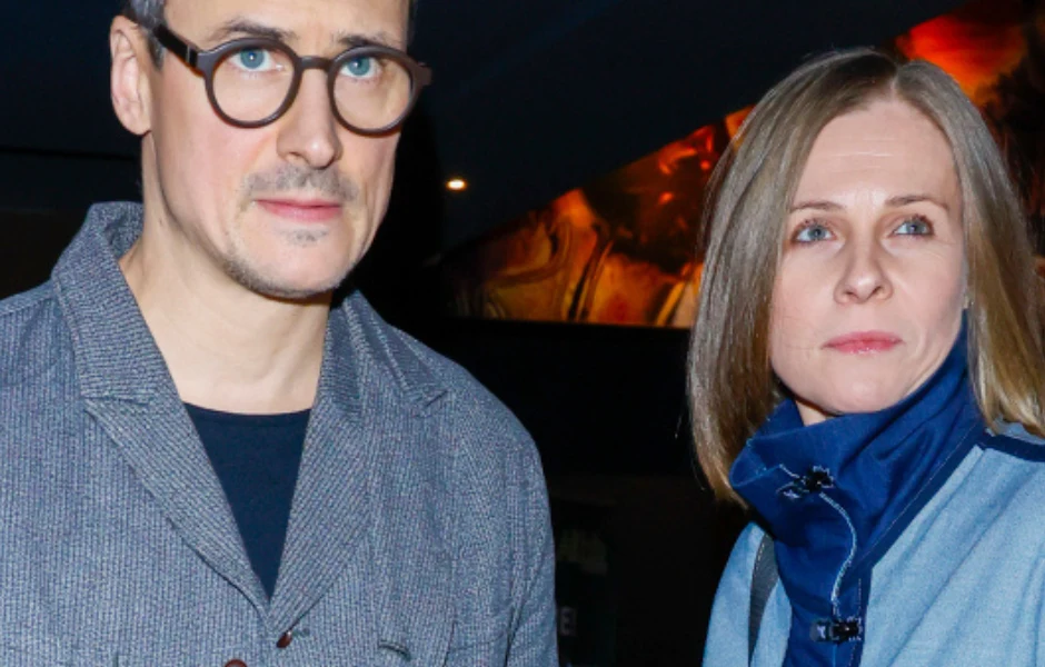 Damięcki szczerze o kryzysie w małżeństwie Dwie dorosłe osoby, kobieta i mężczyzna, stoją obok siebie w średnim planie. Oboje mają neutralny wyraz twarzy, ubrani są w casualowe ubrania w odcieniach szarości i niebieskiego. W tle widoczne rozmyte światło i elementy dekoracyjne.