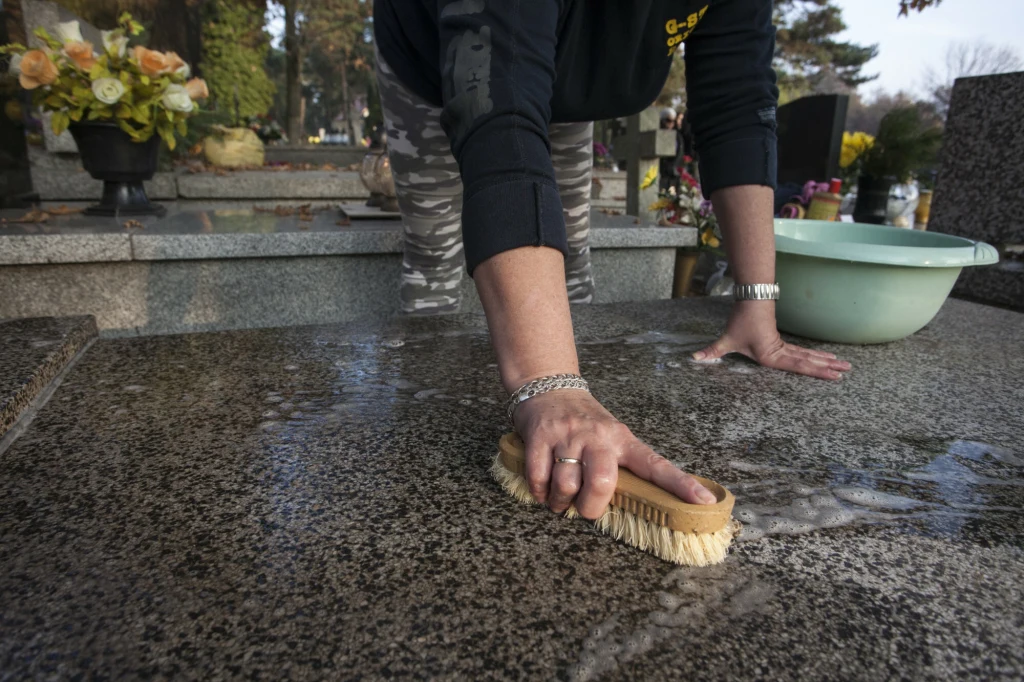 Gorąca woda i płyn do naczyń poradzą sobie z plamami po wosku Czyszczenie pomnika na cmentarzu, ręka osoby trzymającej szczotkę i myjącej nagrobek, w tle widoczne kwiaty i misa z wodą.