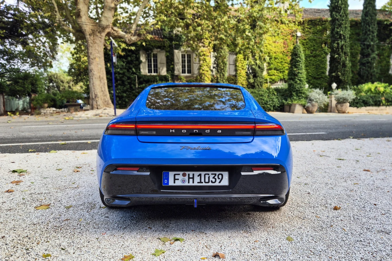 Honda Prelude, jak na hybrydę przystało, ma ukrytą końcówkę wydechu Tył niebieskiego samochodu marki Honda, model z nowoczesnym designem zaparkowany na żwirowym podłożu, w tle drzewa oraz dom pokryty zielonymi pnączami