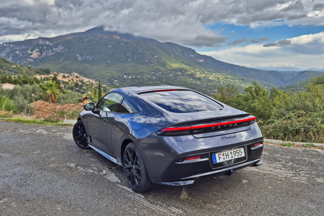 Honda Prelude kosztuje w Polsce 214 500 zł i właściwie nie ma konkurentów Nowoczesny, ciemnoszary samochód sportowy stoi na mokrej nawierzchni, otoczony przez soczyście zielone wzgórza i górzysty krajobraz, na niebie widoczne są chmury zwiastujące deszcz.