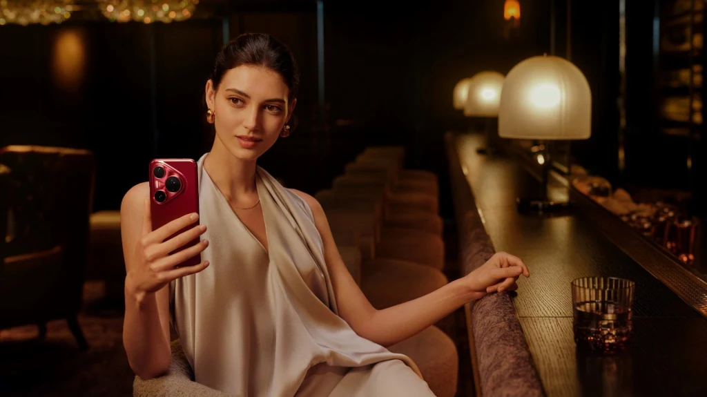 Elegancka kobieta w jasnej sukni siedzi w stylowym, przyciemnionym barze, trzymając w dłoni czerwony smartfon i patrząc w ekran, a na blacie obok stoi szklanka z napojem.
