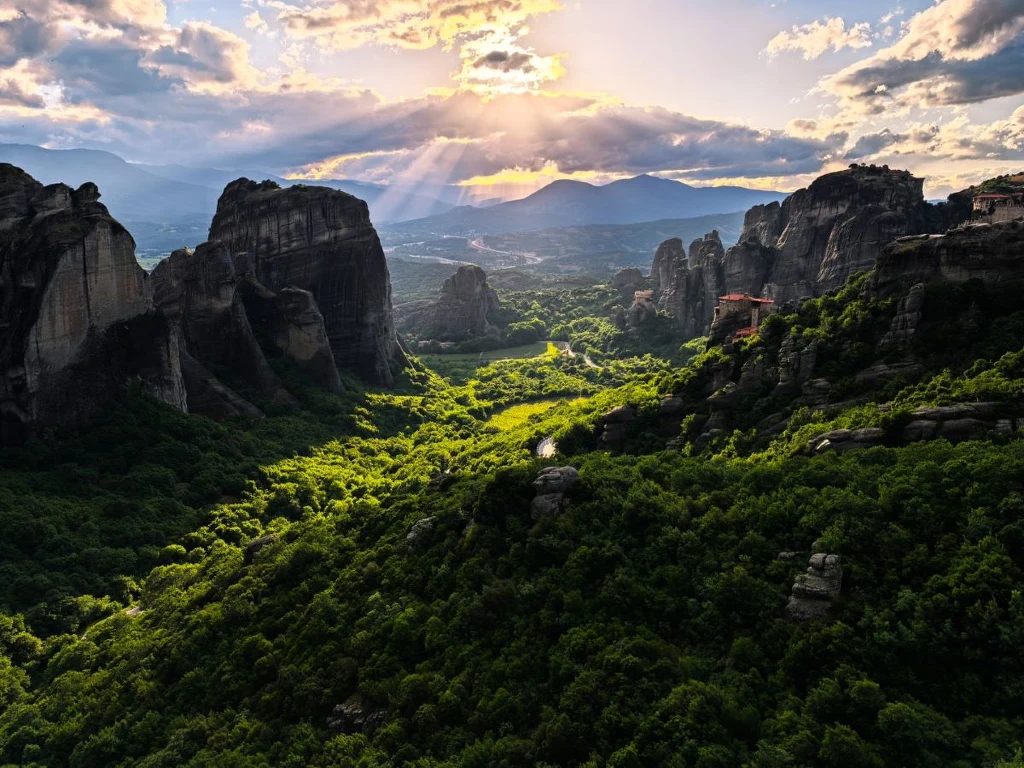 Skały o fantazyjnych kształtach wznoszące się ponad zielonym lasem, pośród których przebijają się promienie słońca, tworząc malowniczy krajobraz górski z klasztorami położonymi na wyżynach.