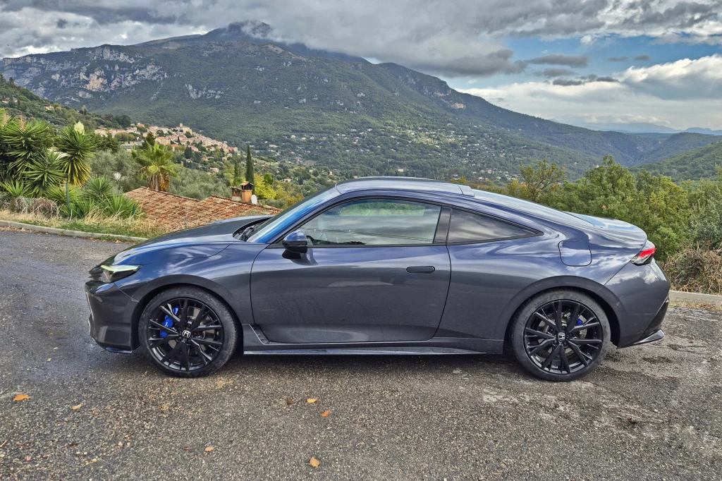 Nowa Honda Prelude ma w sobie sporo charakteru dawnych modeli marki Srebrzystoszary sportowy samochód coupe stoi zaparkowany na poboczu asfaltowej drogi, w tle malowniczy krajobraz górski z zielonymi wzgórzami, chmurami i roślinnością śródziemnomorską.