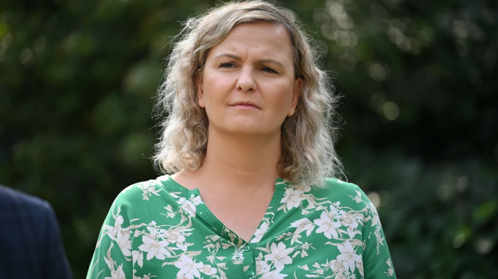 Otylia Jędrzejczak Kobieta o blond kręconych włosach w zielonej bluzce w kwiaty patrzy poważnie przed siebie, w tle rozmazana zieleń ogrodu.