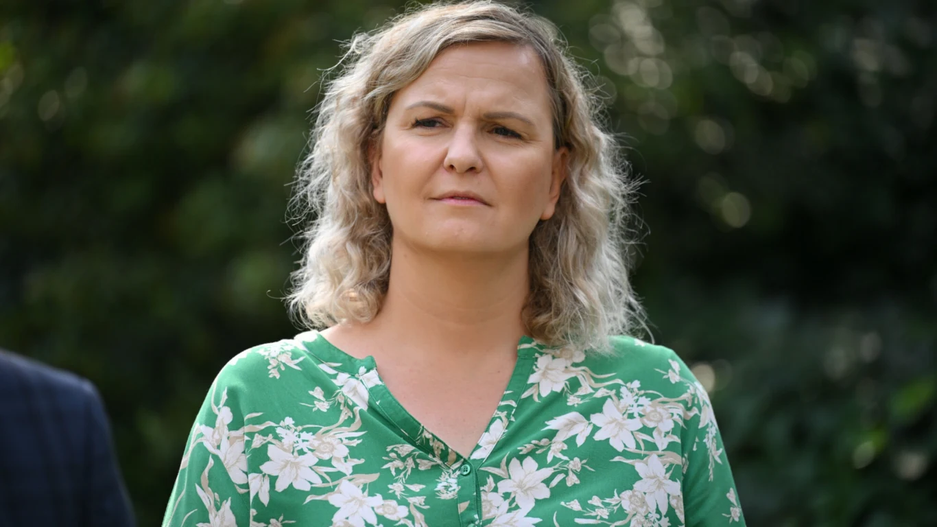 Kobieta o blond kręconych włosach w zielonej bluzce w kwiaty patrzy poważnie przed siebie, w tle rozmazana zieleń ogrodu.