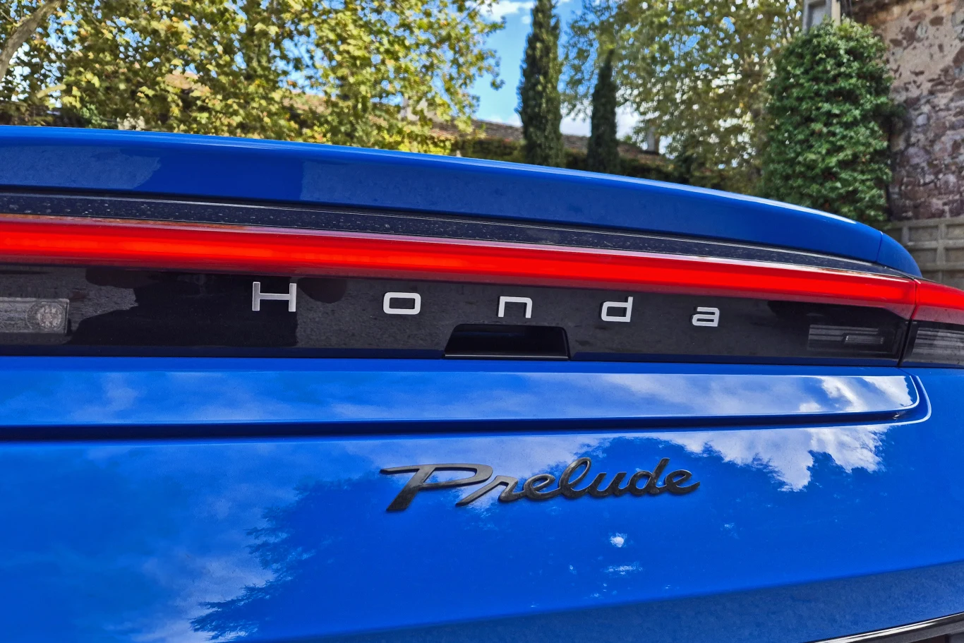 Honda Prelude Tylna część niebieskiego samochodu marki Honda z wyraźnie widocznymi emblematami Honda i Prelude, na tle zielonych drzew oraz fragmentu muru ogrodowego.